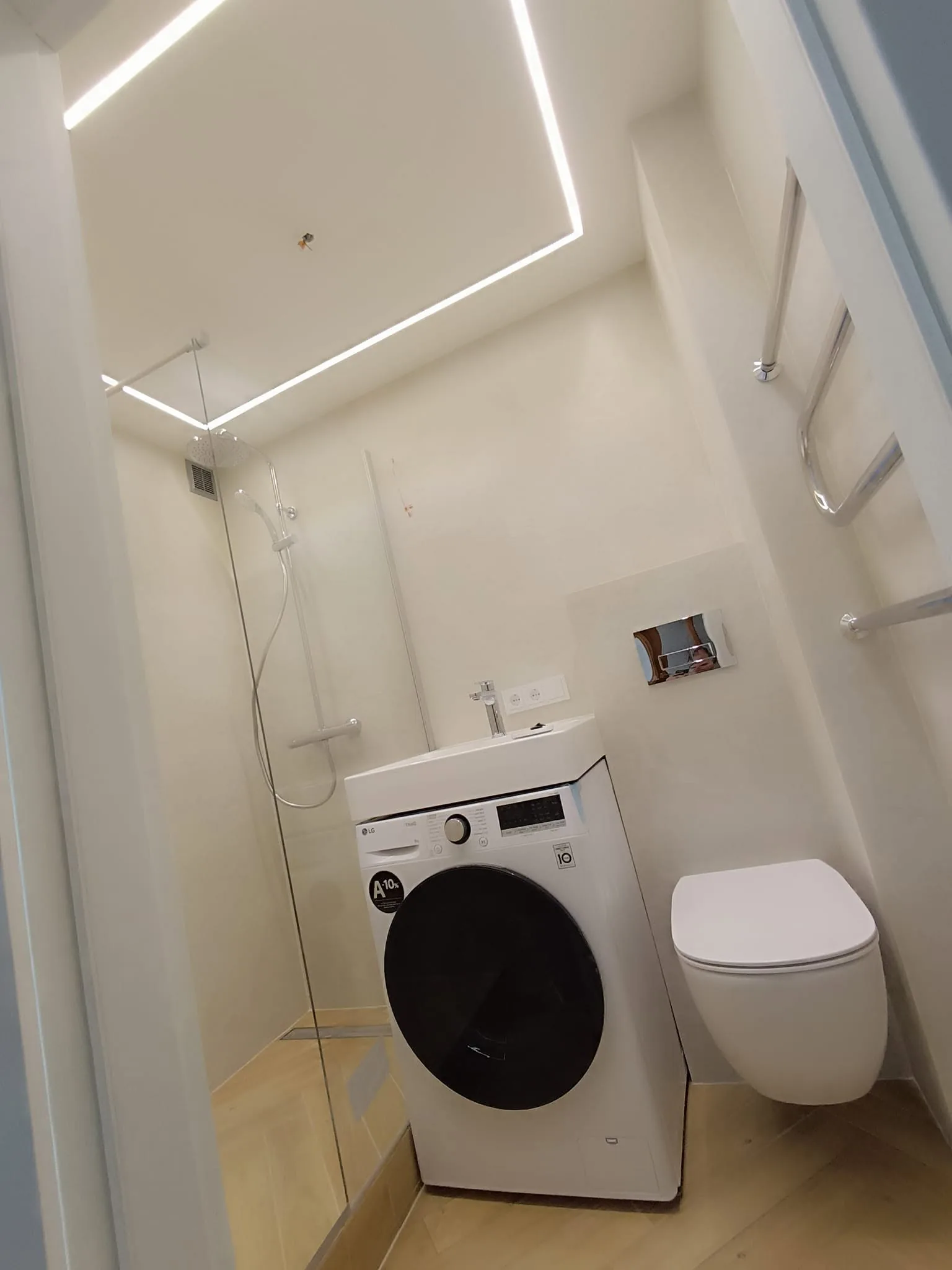 Balta vonios kambario apdaila su LED juostos lubų apšvietimu, stikline dušo pertvara ir pakabinamu WC