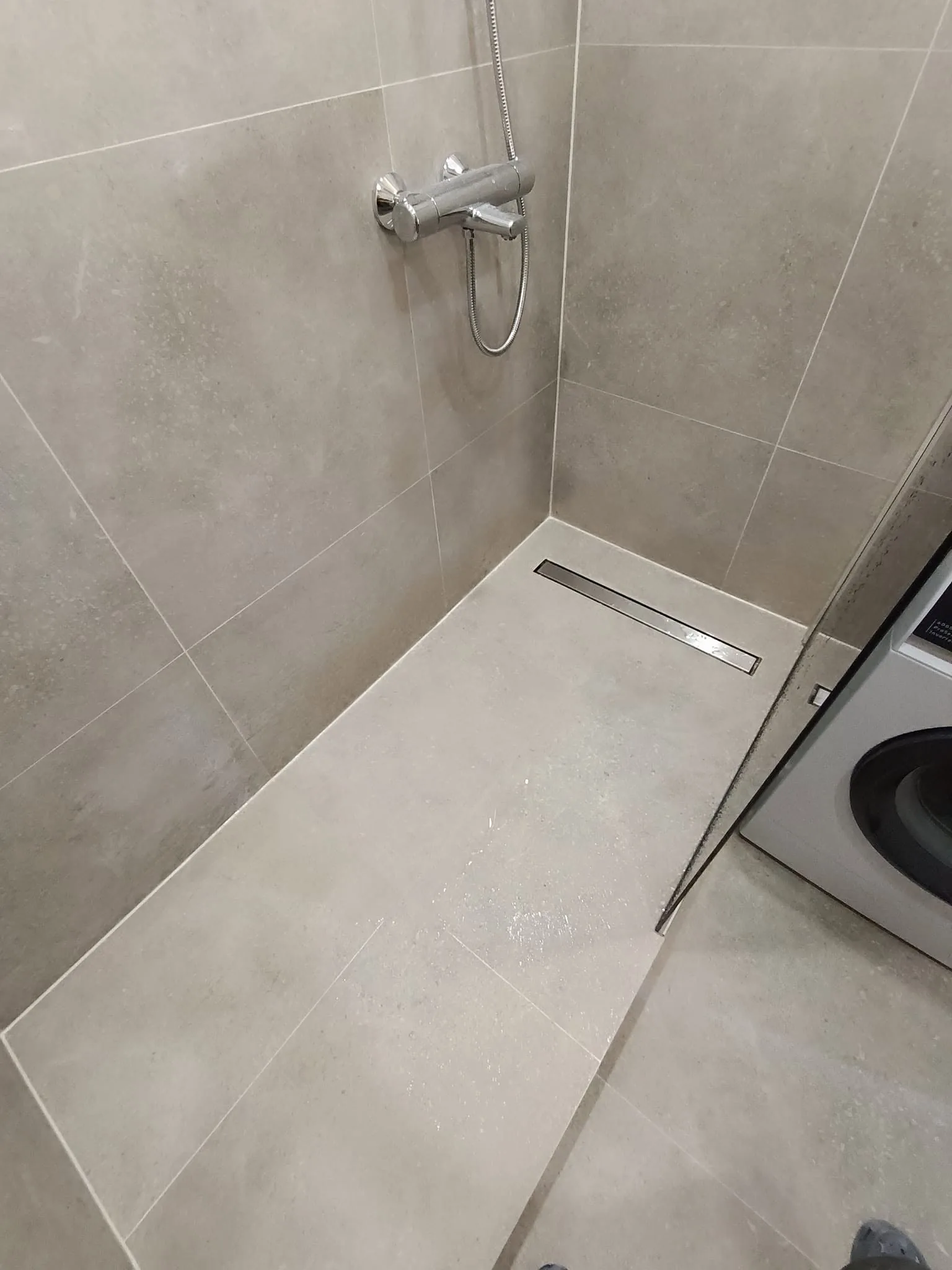 Šviesiomis smėlio spalvos plytelėmis įrengta dušo zona su linijiniu dušo trapu ir stikline pertvara