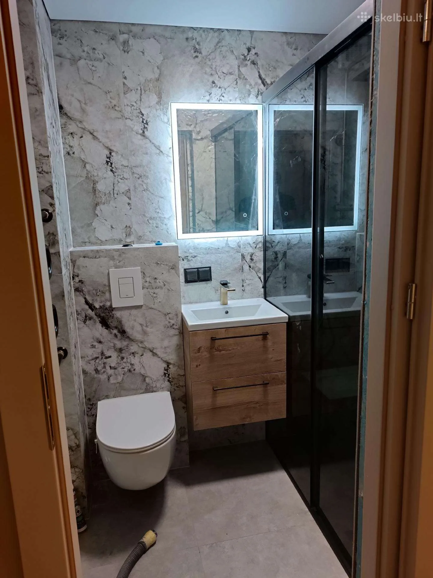 Vonios kambarys su balto marmuro efekto plytelėmis, pakabinamu WC, medinės imitacijos spintelės frontu ir LED veidrodžiu