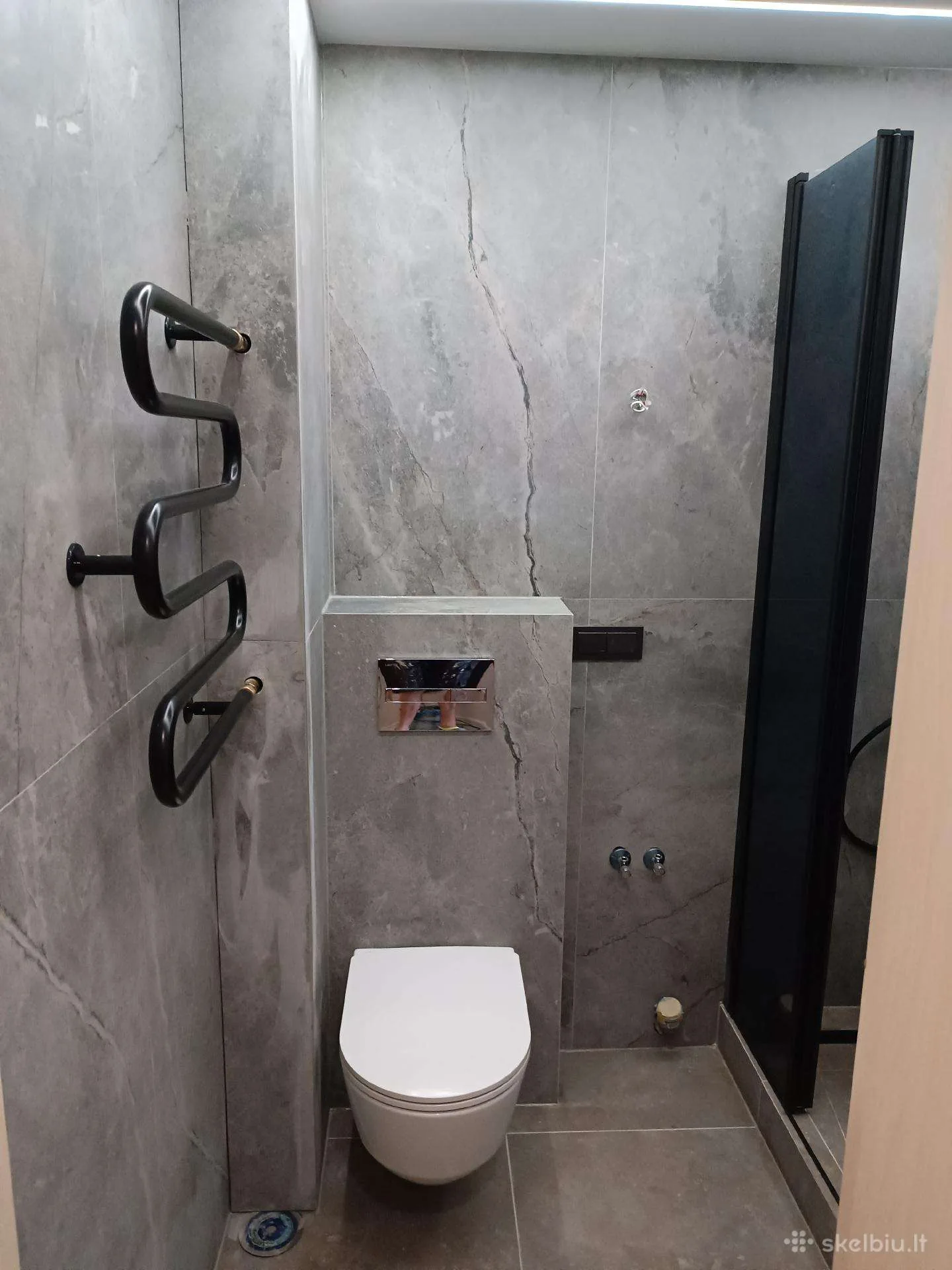 Pilkų marmuro tekstūros plytelių sienos su pakabinamu WC, chromuotu nuleidimo mygtuku ir juodu bangų formos rankšluosčių džiovintuvu